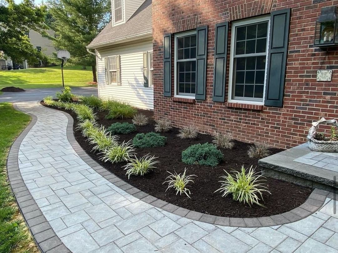 Landscaped walkway with stone path, shrubs, and plants beside a brick house.